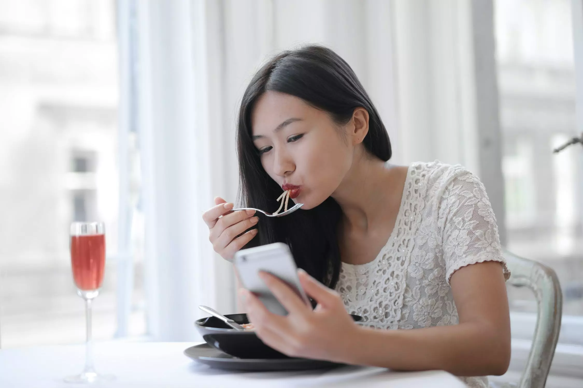 Lady eating a noodle soup dish with a fork texting on her phone